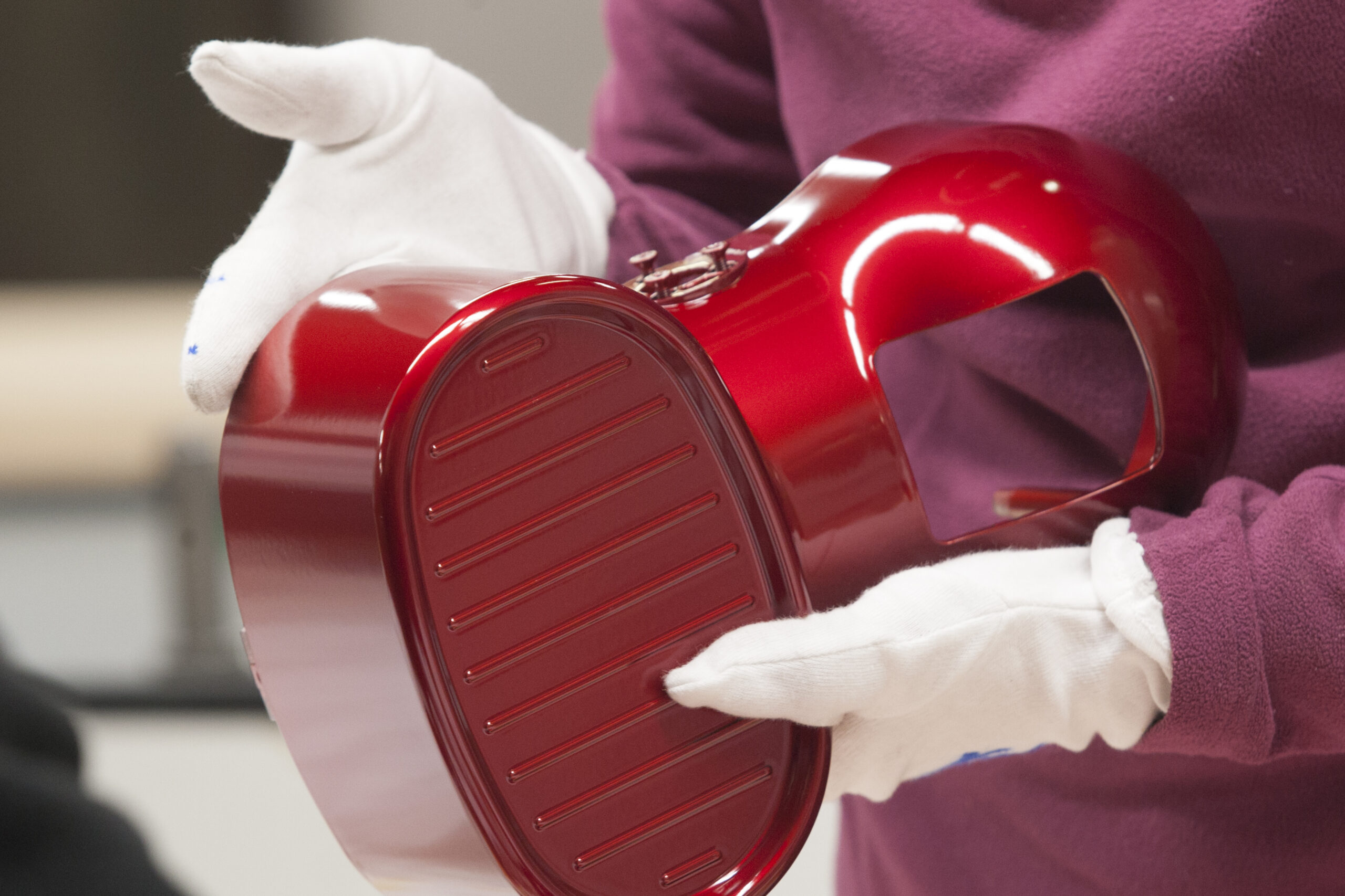 A person wearing white gloves carefully holds a vibrant red die-cast zamak component, showcasing Bruschi's expertise in precision metal casting. The component features a detailed, grooved design.