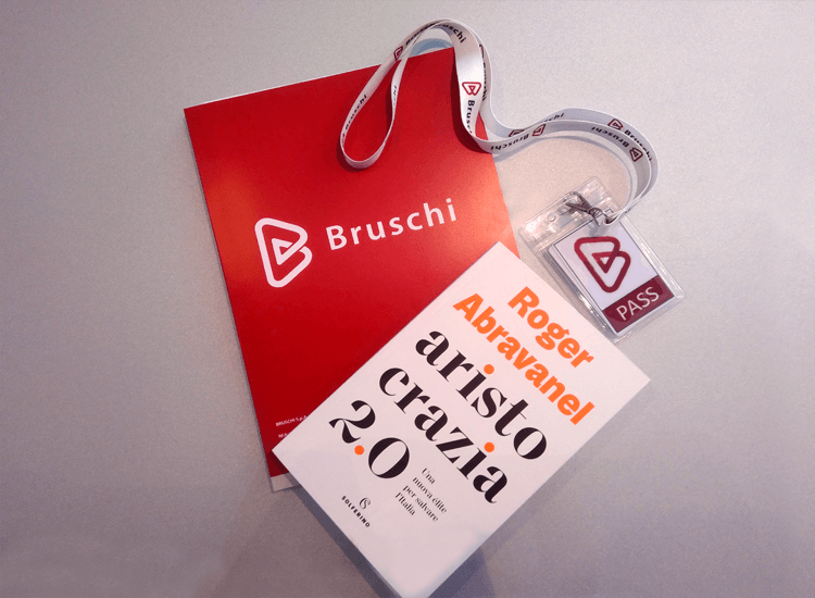 Bruschi company branding materials at an event, showcasing their logo on a red brochure and lanyard. A book, "Aristocrazia 2.0" by Roger Abravanel, is also visible, suggesting a corporate event or partnership.